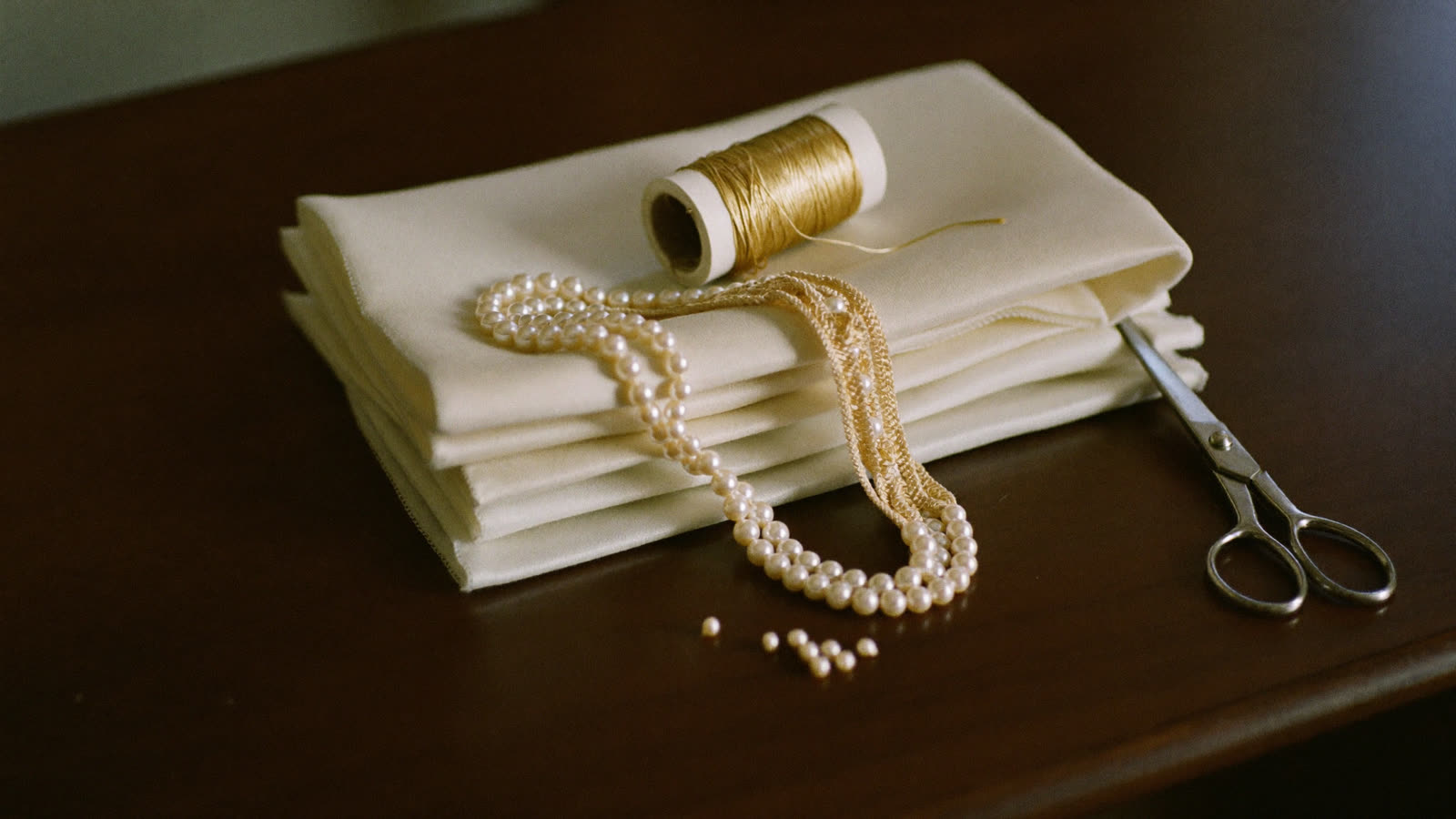 Soies ivoire pliées, bobine de fil doré et perles, sur la table de l’atelier
